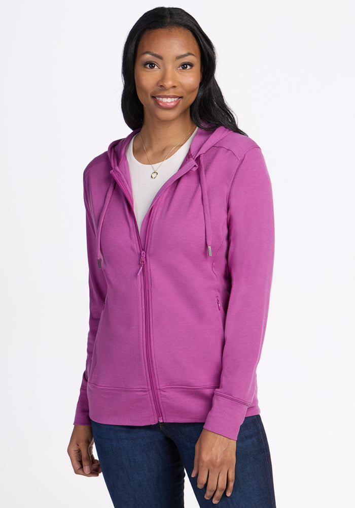 A woman with long dark hair is smiling, wearing the Woolx Ryann Hoodie in Purple Wine over a white shirt and blue jeans, standing against a plain white background. 