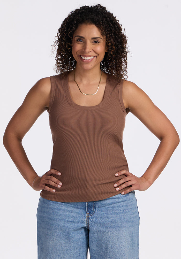 A woman with curly hair smiles at the camera, standing with her hands on her hips. She is wearing the Woolx Sloane Ribbed Tank in Sienna Kiss and blue jeans against a plain white background. 