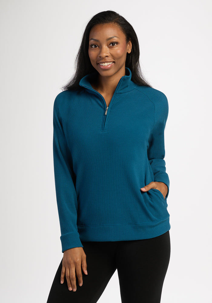 Woman wearing Woolx Quinn Ribbed 1/4 Zip in Blue Pine and black leggings, smiling with a hand in her pocket. 