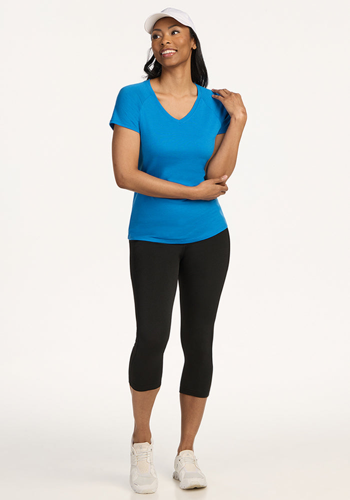 A woman wearing the Woolx Mia V Neck - Wave Rider, a blue moisture-wicking women’s v-neck t-shirt, with a white cap, black capri leggings, and white sneakers stands smiling with one hand on her shoulder against a plain light background.