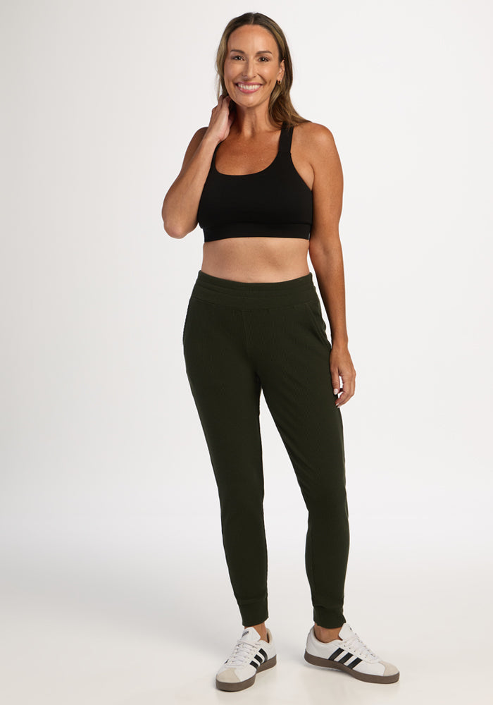 Woman smiling, wearing Woolx Lennon Ribbed Joggers in Duffel Bag, a black sports bra, and white sneakers.