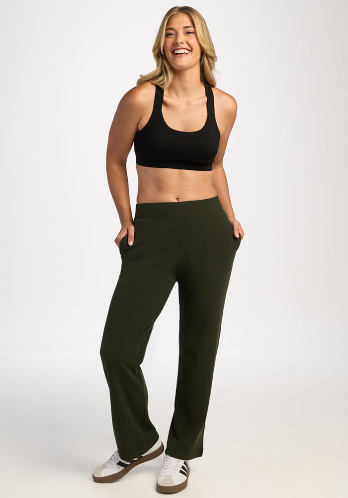 Smiling woman in black sports bra and Woolx Luca Petite Pants - Duffel Bag stands hands-in-pockets, white background.