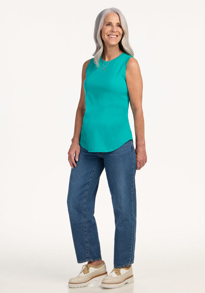 A smiling older woman with long gray hair wears the Woolx Bella Tunic Top in Cool Jade, paired with blue jeans and beige platform shoes, standing against a plain white background.
