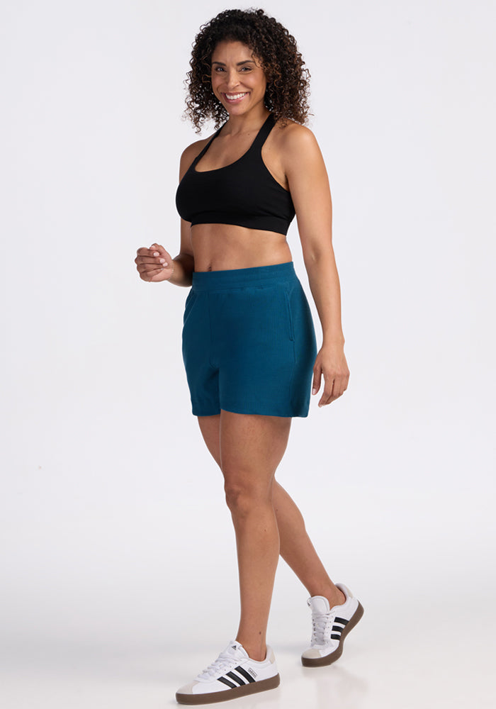 A woman with curly hair smiles while posing in a black sports bra, Woolx Maisey Ribbed Shorts in Moroccan Blue, and white Adidas sneakers against a plain white background.