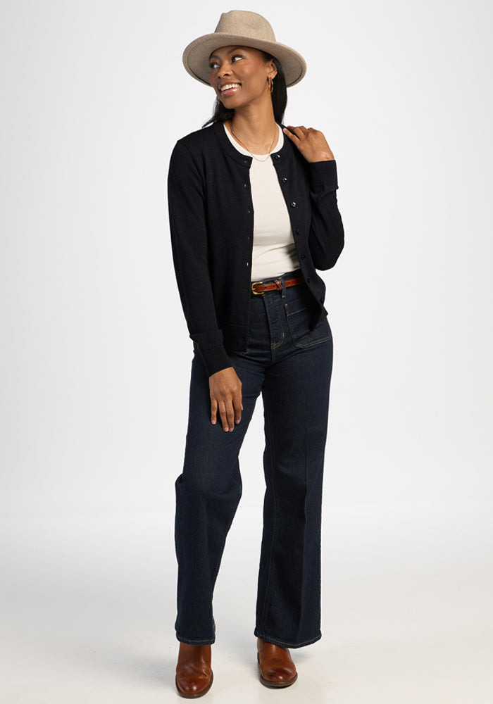 Woman in Caitlin Sweater - Black by Woolx, white shirt, jeans, boots, and hat stands smiling against plain background.