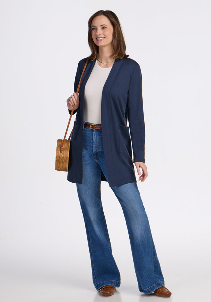 A woman smiles against a white background, wearing the Woolx Paisley Cardigan in Deep Navy with a white top, flared blue jeans, brown shoes, and holding a tan woven crossbody bag.