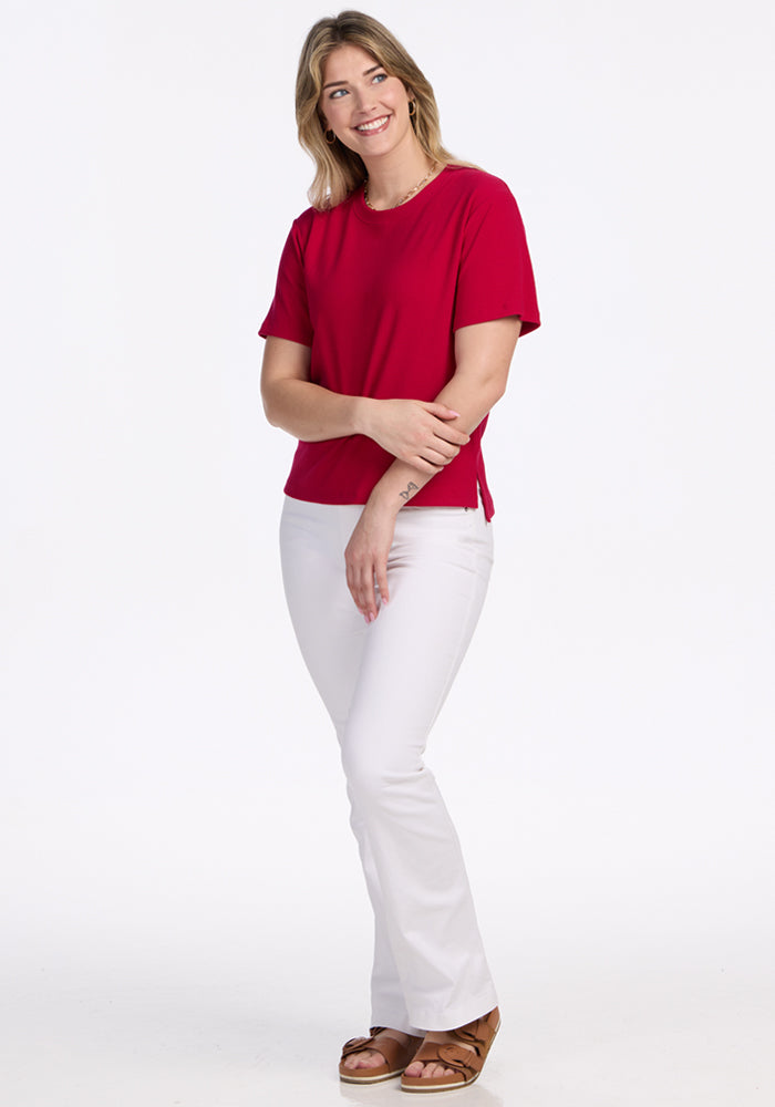 A woman with light hair stands smiling against a white background, arms gently crossed, wearing the Woolx Delia Ribbed Short Sleeve in Poppy Spark, white pants, and brown sandals.