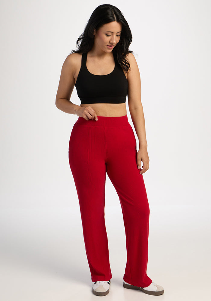 Woman in a black sports bra and Woolx Luca Ribbed Pants in Maraschino Cherry stands looking down, hand on waistband.