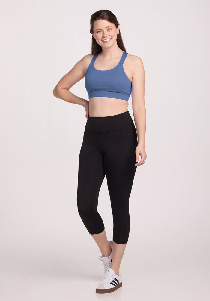 A woman smiles, wearing the Woolx Livi Bra - Skyline, paired with black cropped leggings and white sneakers with black stripes. She poses confidently against a plain light background, with one hand on her hip.