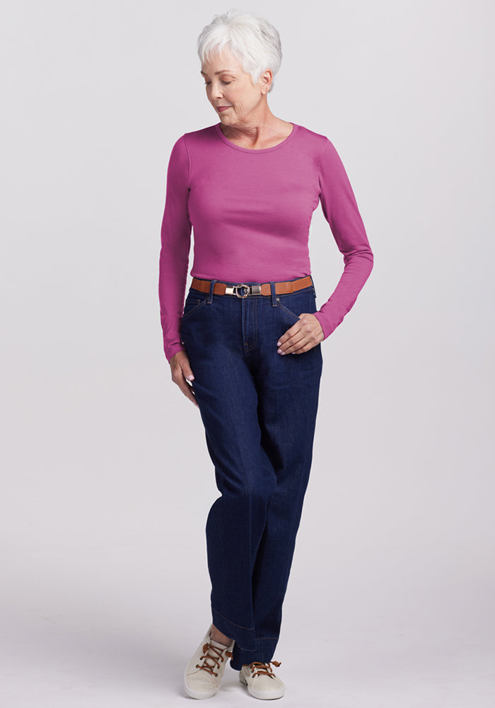 Older woman with short white hair wears the Woolx Remi Long Sleeve T-Shirt in Purple Wine, blue jeans, brown belt, and white sneakers. She stands with one leg crossed, looking down against a plain light background. The shirt is odor-resistant and moisture-wicking.