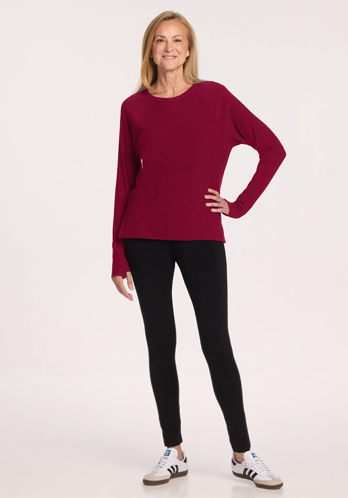 Woman wearing the Woolx Ainsley Sweater in Beet Red, black leggings, and white sneakers, smiling at the camera.