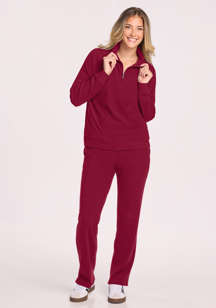 Smiling person in a Woolx Quinn Ribbed 1/4 Zip - Beet Red, pants, and white sneakers against a plain background.