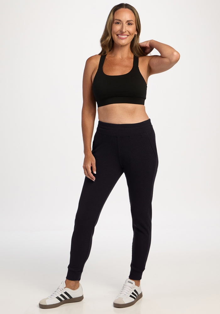 Smiling woman in black Woolx Lennon Ribbed Joggers, black sports bra, and white sneakers, hand on head.