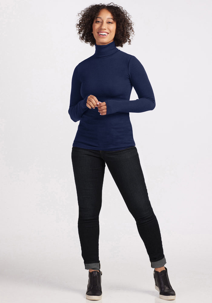 Woman smiles in a Woolx Sage Ribbed Turtleneck - Ocean Cavern, black jeans, and sneakers against a light backdrop.