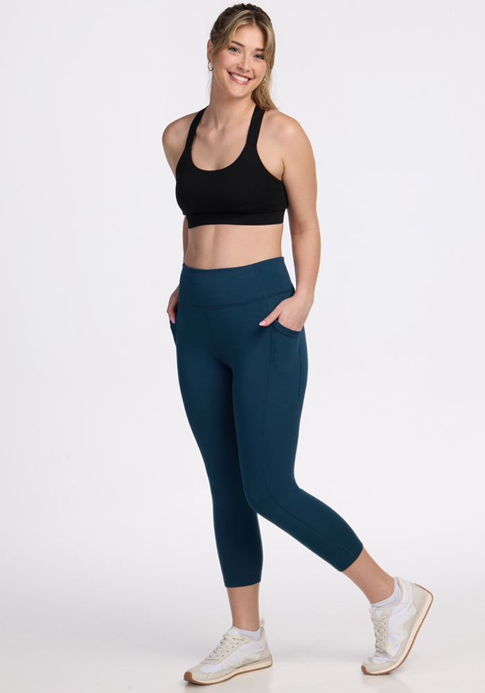 A woman stands smiling with her hands in her pockets, wearing a black sports bra, white sneakers, and Woolx Merino Flex® McKenna Capris in Majolica Blue against a plain white background.