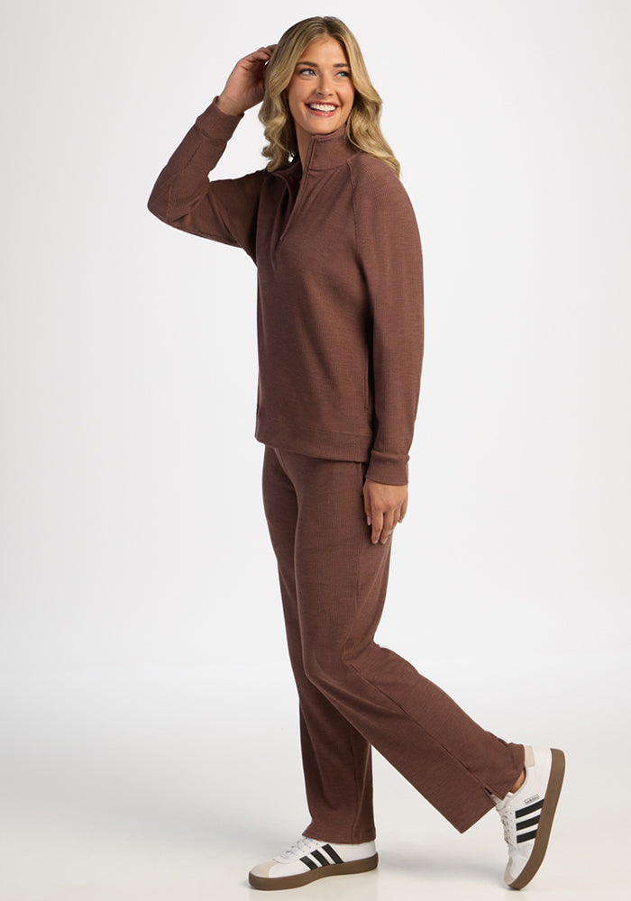 Woman in Woolx Quinn Ribbed 1/4 Zip - Sable Heather and white sneakers smiles, hand in hair, on white backdrop.