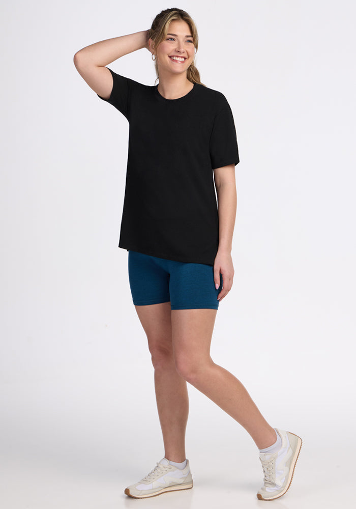 A smiling person stands with one hand behind their head, wearing the Woolx Audrey Tee in black, blue shorts, and white sneakers against a plain white background.