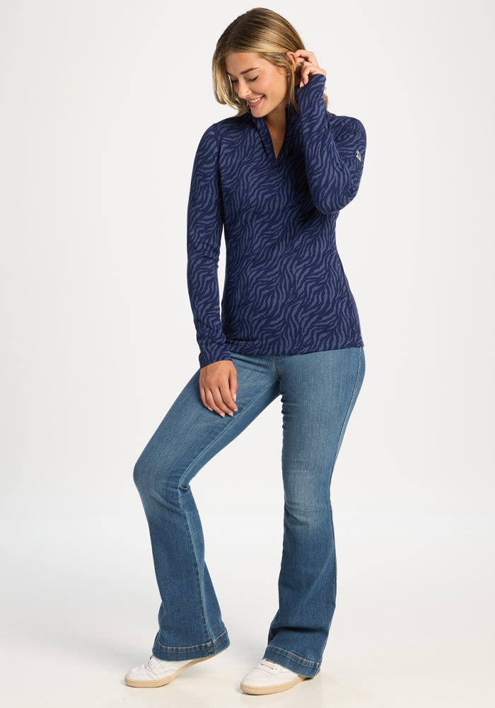 Smiling woman in Woolx Emi 1/4 Zip - Midnight Safari and blue jeans stands with one knee bent, looking down.
