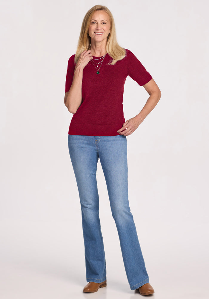Woman wearing Woolx Anna Crew Sweater in Beet Red, blue jeans, and brown shoes standing against a white background.