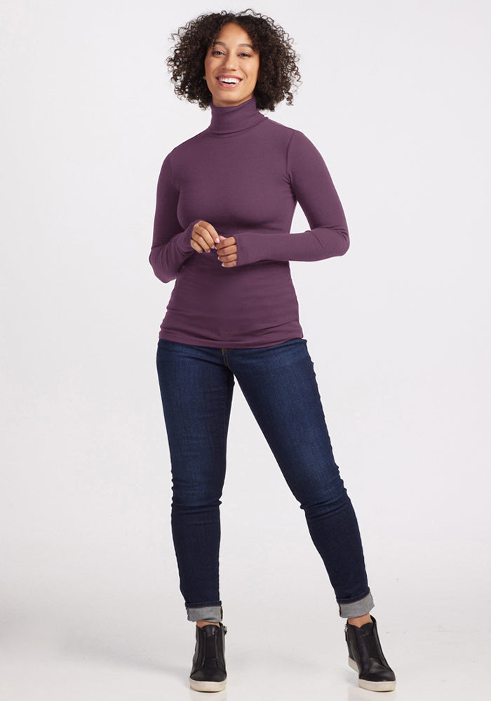 Woman smiling in a Woolx Sage Ribbed Turtleneck - Purple Iris, dark jeans, and black sneakers against a light background.