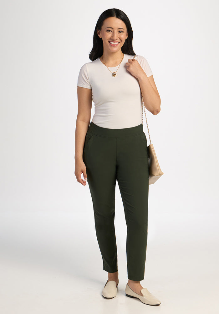Woman wears Woolx Nicole Tall Ankle Pant in Duffel Bag and a white tee, smiling, holding a beige purse.