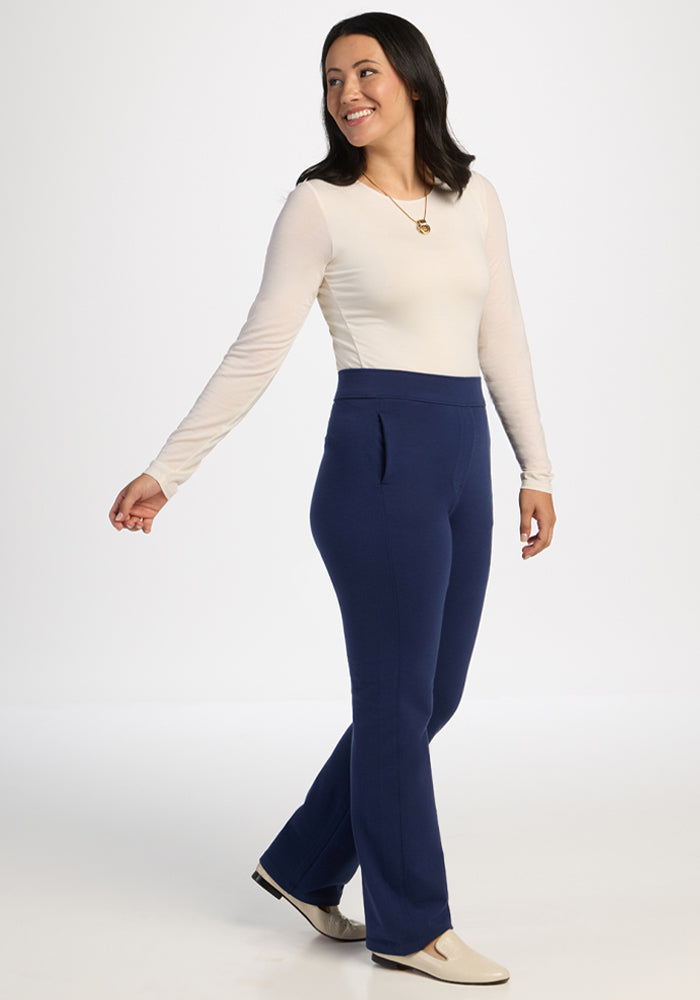 Smiling woman in a cream top and Woolx Emerson Straight Leg Pants Petite - Ocean Cavern walks by a white background.