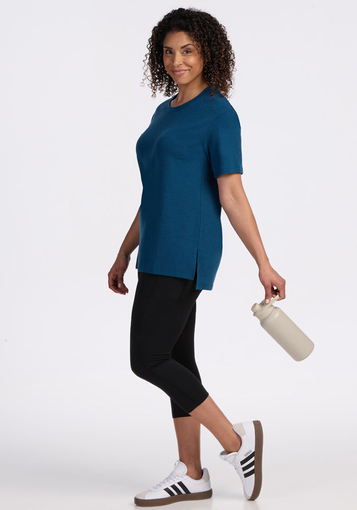 A woman with curly hair wears the Woolx Audrey Tee in Majolica Blue, paired with black leggings and white sneakers. She smiles, holding a beige water bottle while walking against a plain white background.