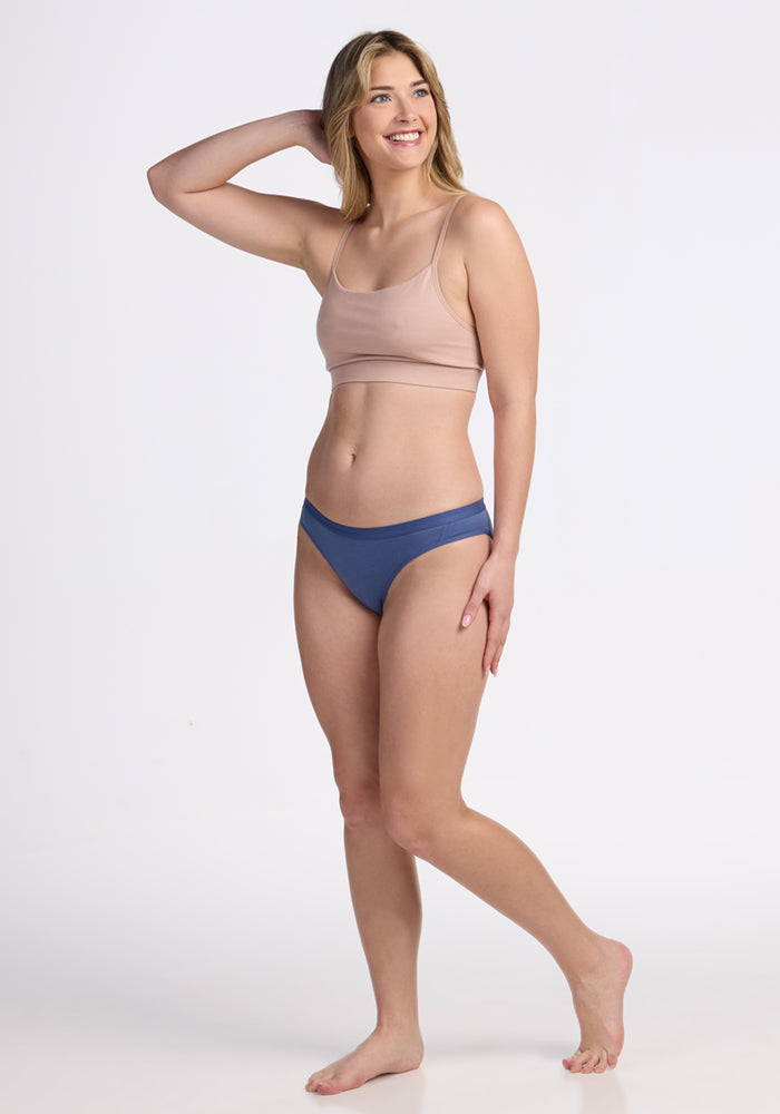A woman in a light beige crop top and blue Woolx Roxie Bikini Underwear - Skyline, made from moisture-wicking Merino wool, stands barefoot against a plain white background, smiling and looking to the side with one hand touching her hair.
