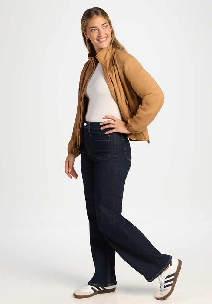 A woman wears a tan, all-season Cooper Windbreaker - Saddleback, a white top, dark wash jeans, and sneakers.