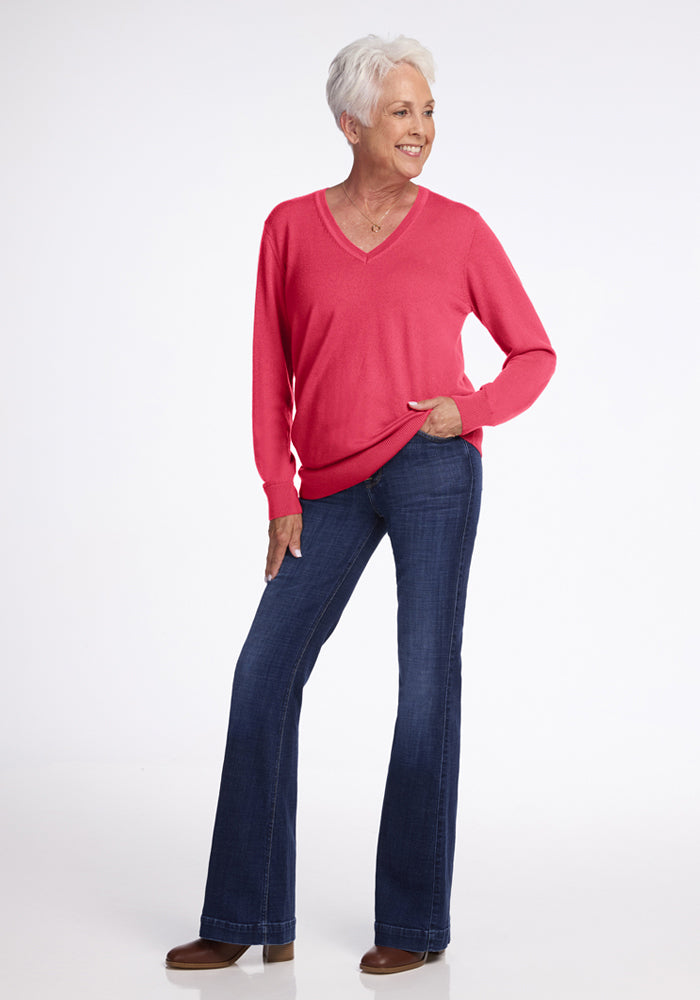 Smiling older woman wears the Woolx Francesca Sweater in Vivid Rose with blue jeans, hand in pocket, on white backdrop.