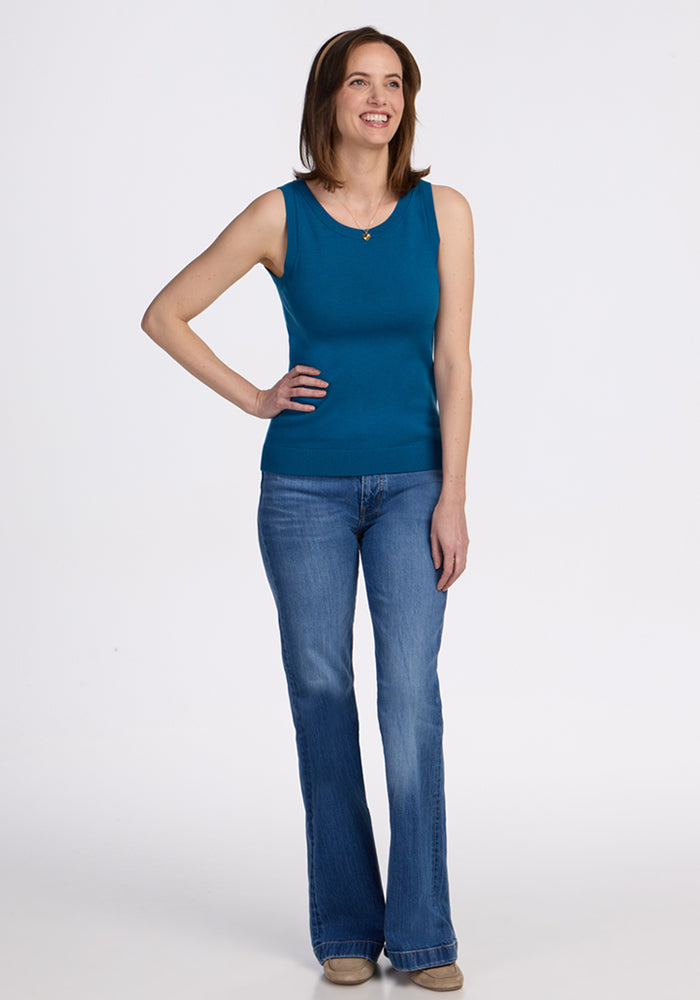 A smiling woman with shoulder-length brown hair stands on a white background, wearing the Woolx Chessy Tank Top in Moroccan Blue as a stylish layering piece, paired with blue jeans and beige shoes. Her right hand rests on her hip.