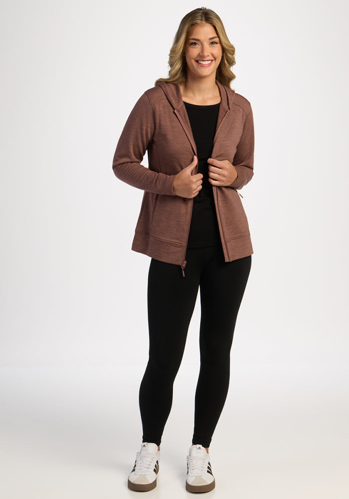 Woman in Woolx Darby Zip Up Hoodie - Sable Heather, black shirt, leggings & white sneakers smiles against a white background.