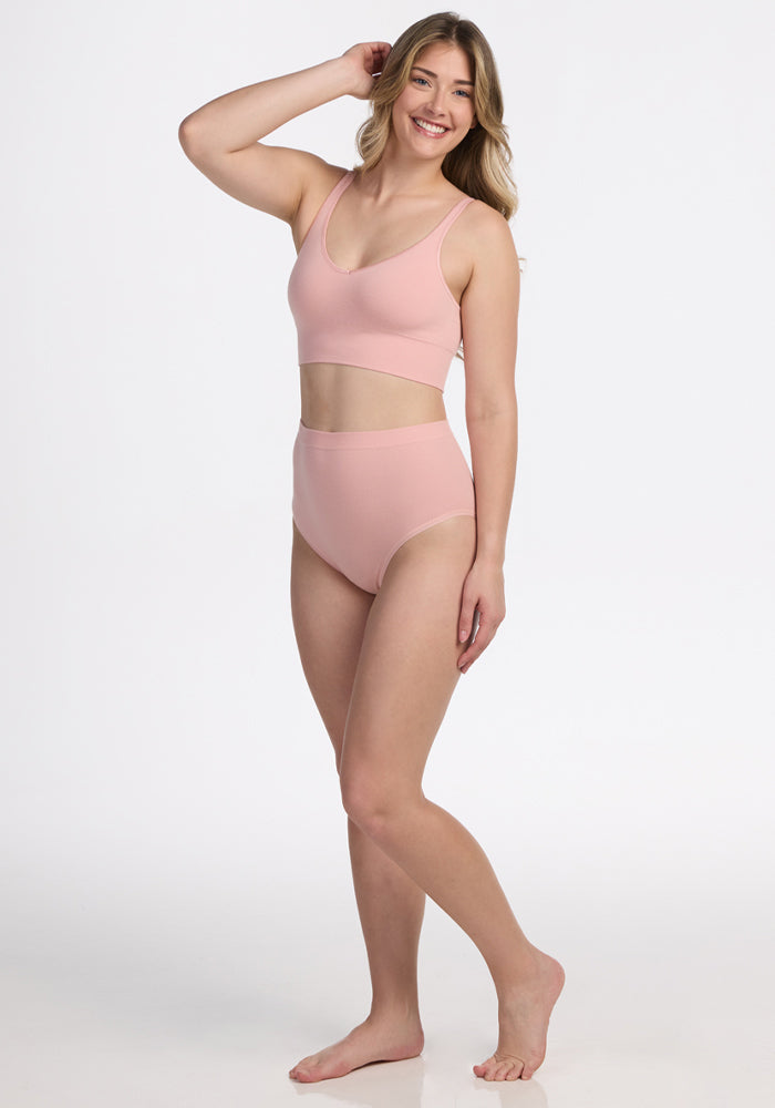 A woman with long blonde hair poses barefoot, smiling in Woolx’s Mabel Full Coverage Underwear in Rosewater and a matching sports bra, set against a plain white background. 