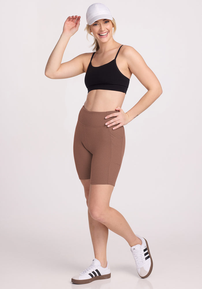 A smiling woman poses against a plain light background in a black sports bra, Woolx Miley Bike Shorts in Sienna Kiss, a white cap, and white sneakers, with one hand on her hip and the other adjusting her hat.