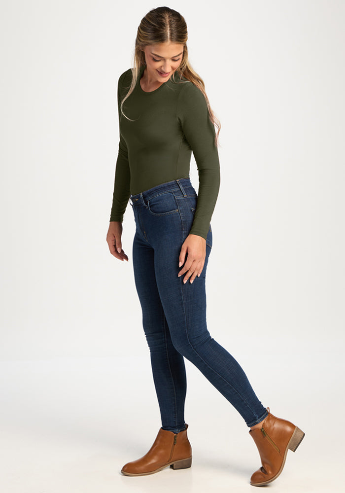 Woman in a Woolx Margot Bodysuit - Everleaf, blue jeans, and brown boots stands looking down and smiling.