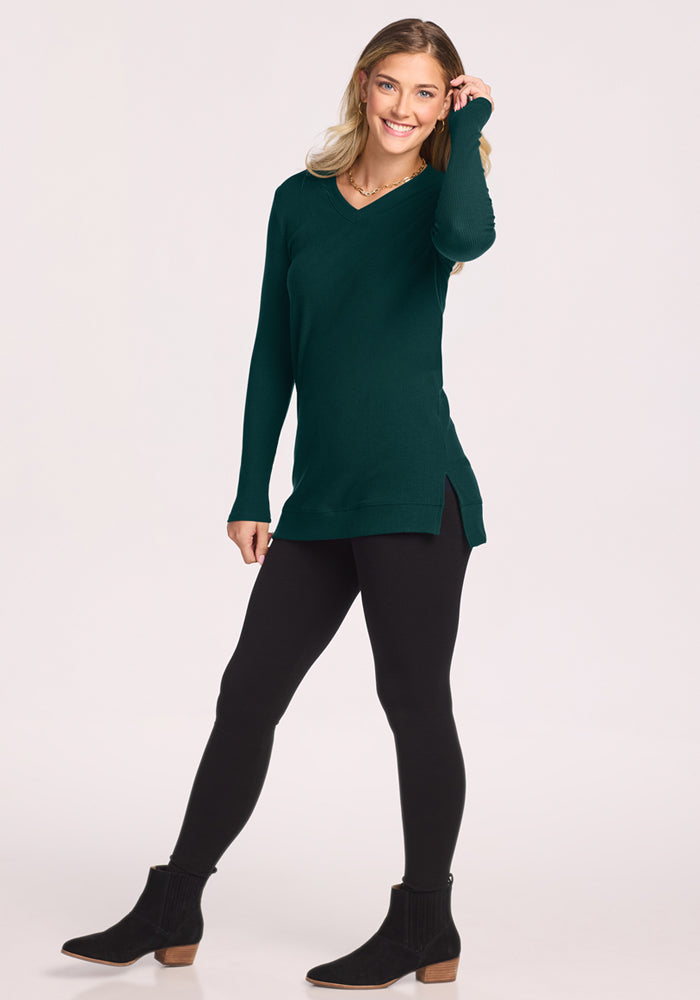 Woman in the Woolx Evie Ribbed Tunic - Deep Pine, black leggings, and boots smiles with one hand in her hair.