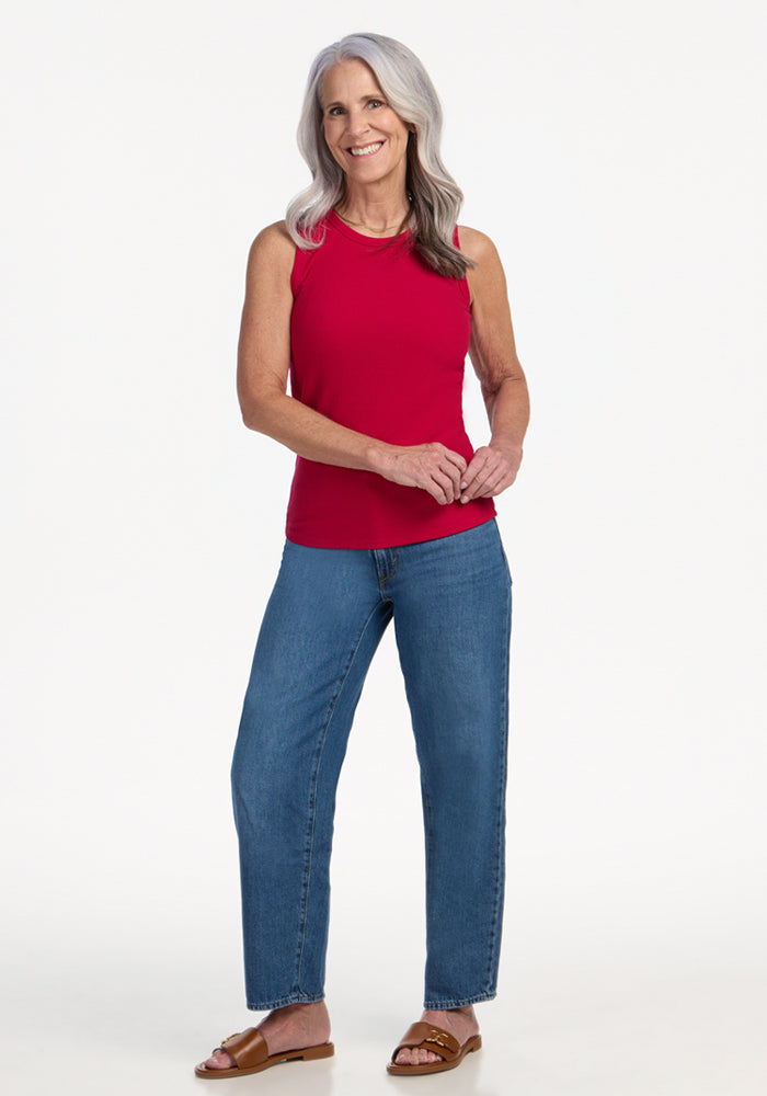 A smiling older woman with long gray hair wears the Woolx Raegan Ribbed Tank Top in Poppy Spark, styled with blue jeans and brown sandals, as she stands against a plain white background.