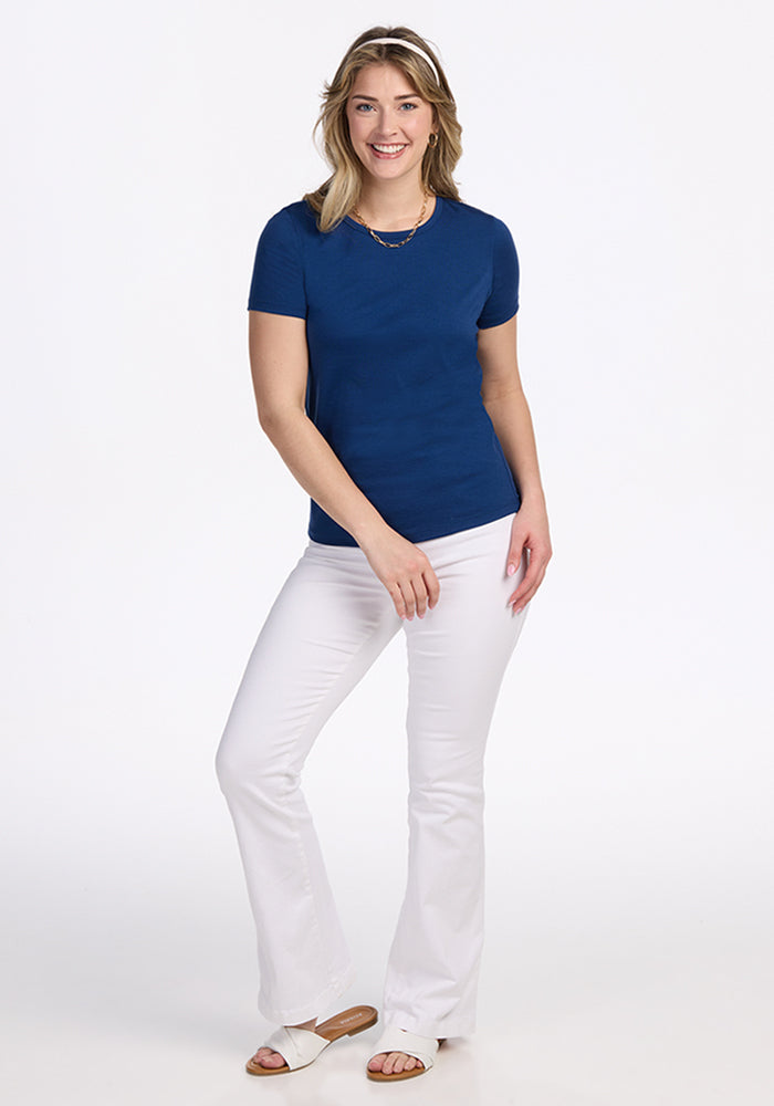 A woman with light brown hair smiles against a white background, wearing the Woolx Liza Crew Neck Tee in Estate Blue, paired with white flare pants, white sandals, and a necklace. Her hands rest at her sides.