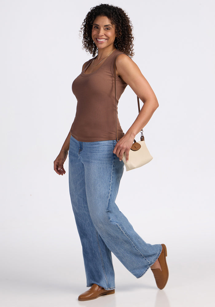A woman with curly hair smiles, balancing on one leg, wearing the Woolx Sloane Ribbed Tank in Sienna Kiss, blue wide-leg jeans, brown shoes, and a small beige purse. She stands against a plain white background.