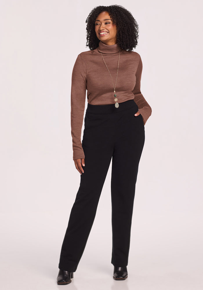 Woman in the Woolx Peyton Turtleneck - Sable Heather, black pants, and boots, smiling with a hand in her pocket.