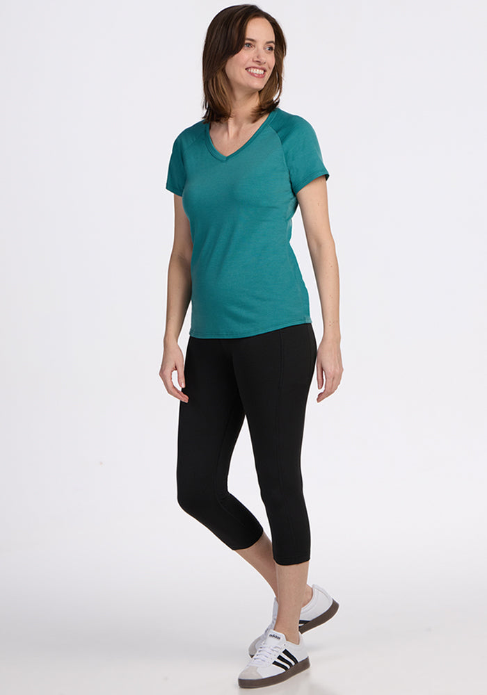 A woman wearing a teal Woolx Mia V Neck - Surfside Merino wool tee with short sleeves, black capri leggings, and white Adidas sneakers stands and smiles against a plain white background.