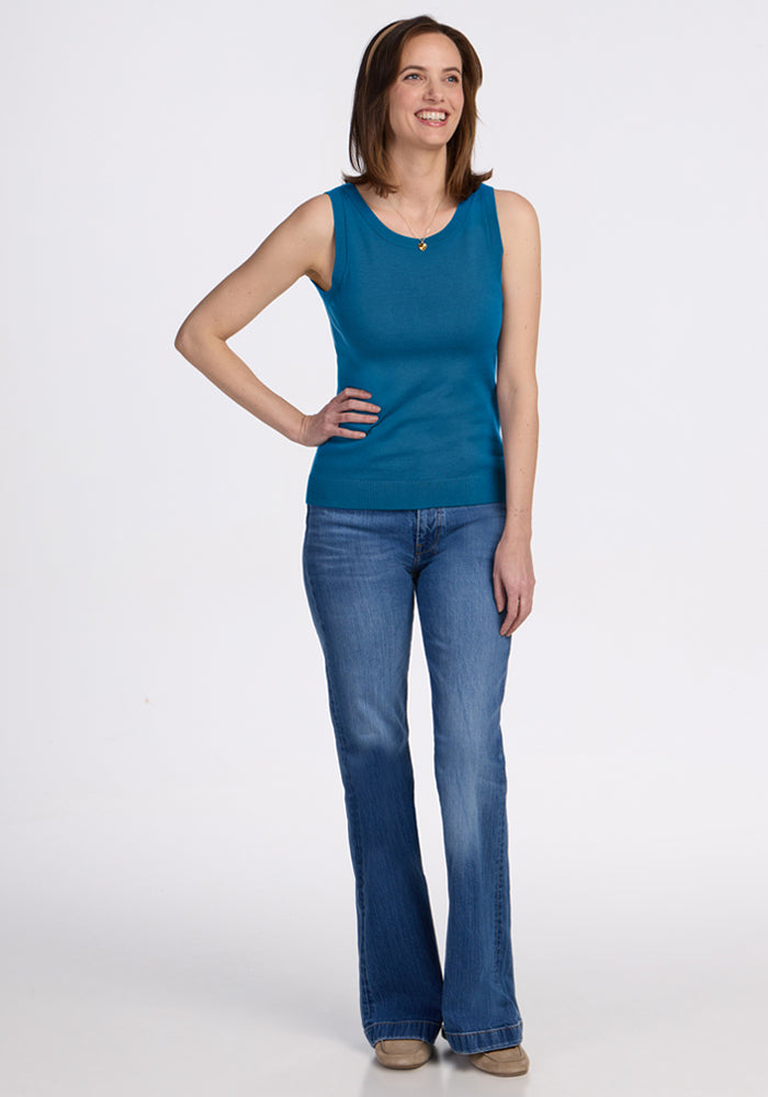 Woman wears Woolx Chessy Tank Top - Blue Pine and jeans, smiling with a hand on her hip against a white background.