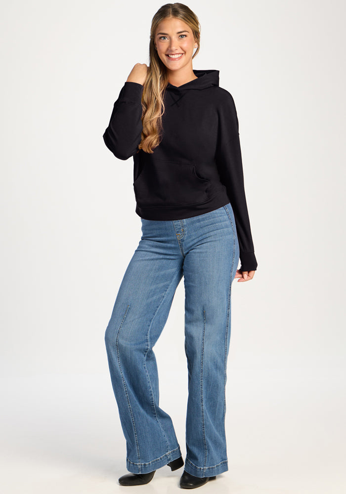 Woman in the Woolx Lizzy Hoodie - Black and blue jeans smiles, hand in hair, against a plain background.