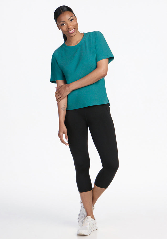 A woman smiles with her arms crossed, wearing the Woolx Delia Ribbed Short Sleeve in Surfside, black cropped leggings, and white sneakers against a plain white background.