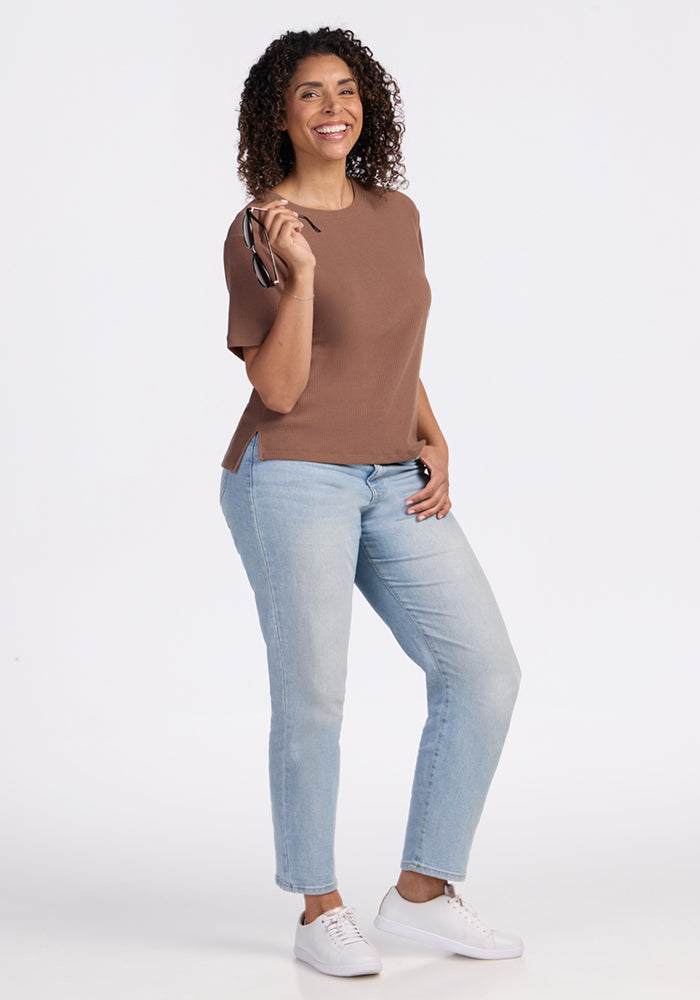A smiling woman with curly hair holds eyeglasses, wearing the Woolx Delia Ribbed Short Sleeve in Sienna Kiss, light blue jeans, and white sneakers against a plain white background.