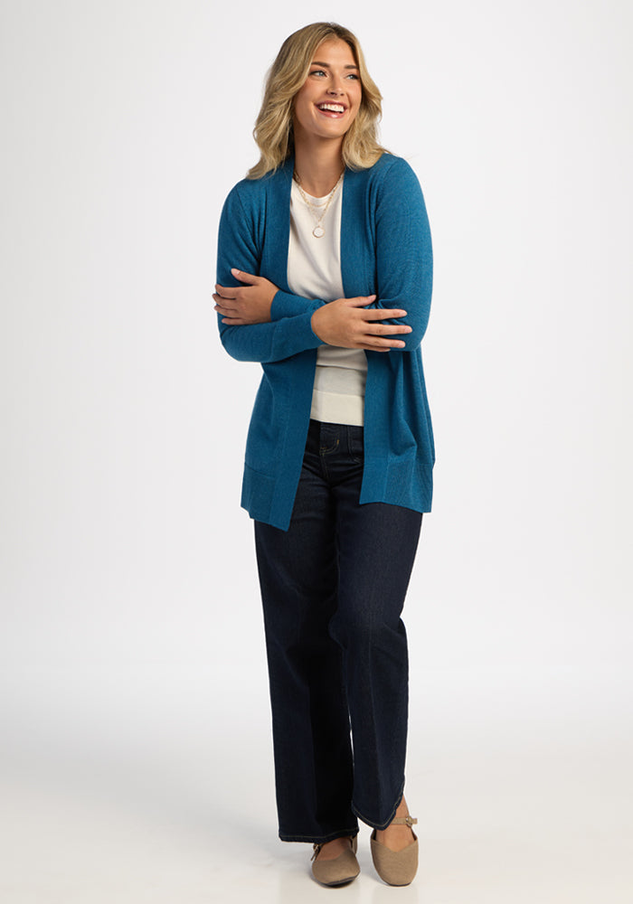 Smiling woman wears the Woolx Macie Sweater in Blue Pine, white shirt, and dark jeans against a plain background.