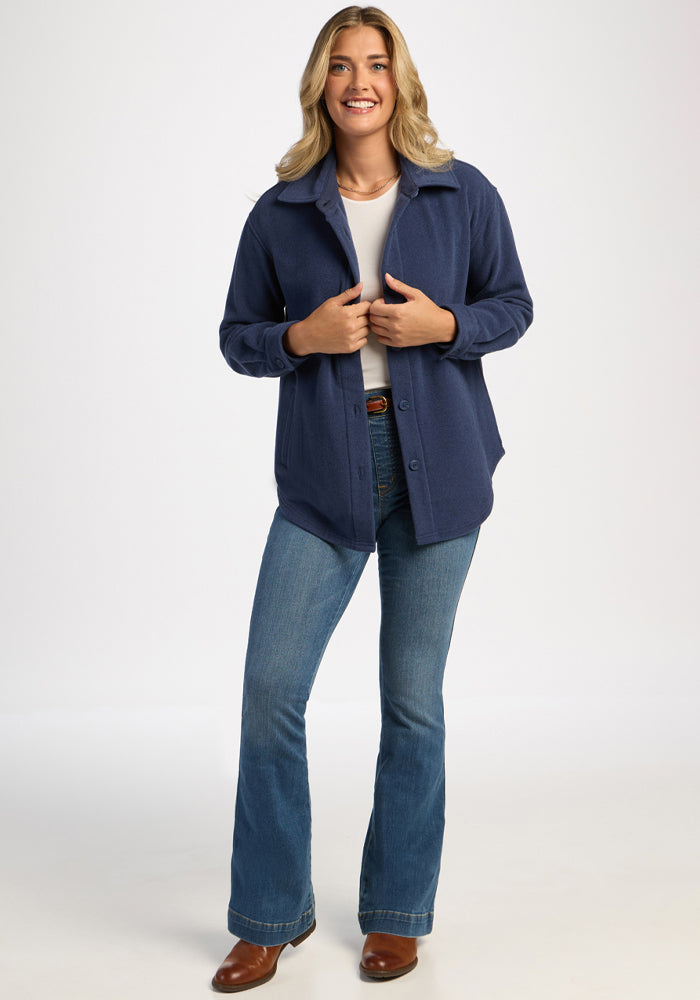 Smiling person in the Woolx Jovie Shacket - Deep Dive, white shirt, jeans, and brown shoes against a plain background.