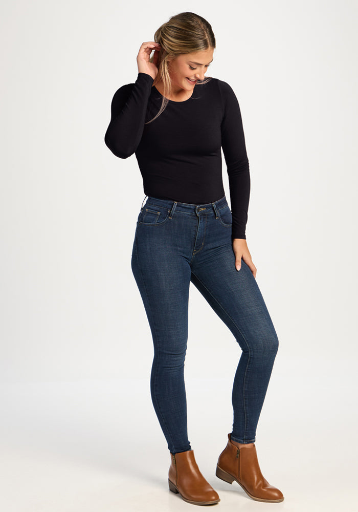 Woman in Woolx Margot Bodysuit - Black, blue jeans, and brown boots stands smiling, looking down.