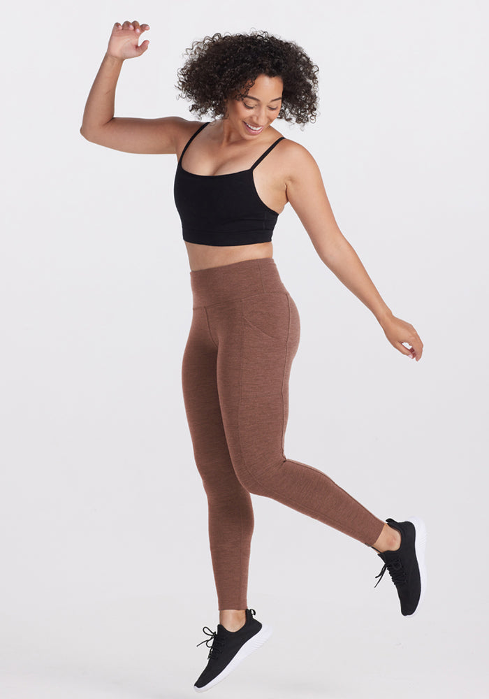 Woman in Woolx Piper Pocket Leggings - Sable Heather jumps joyfully on one foot against a plain white background.