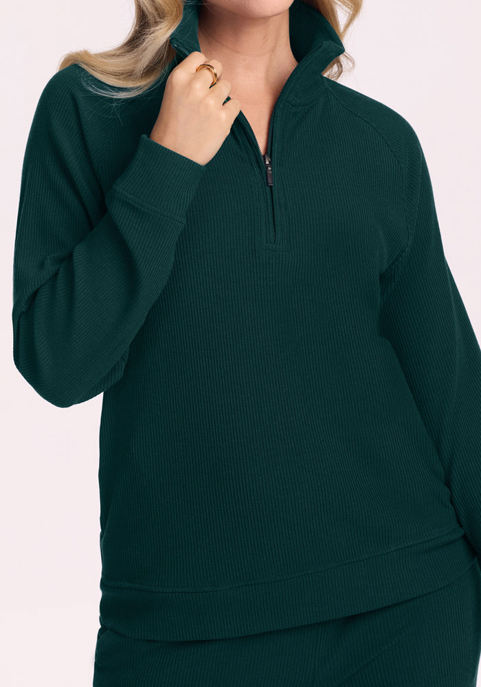 Woman in a Woolx Quinn Ribbed 1/4 Zip - Deep Pine, holding the collar, wearing gold rings, light background.